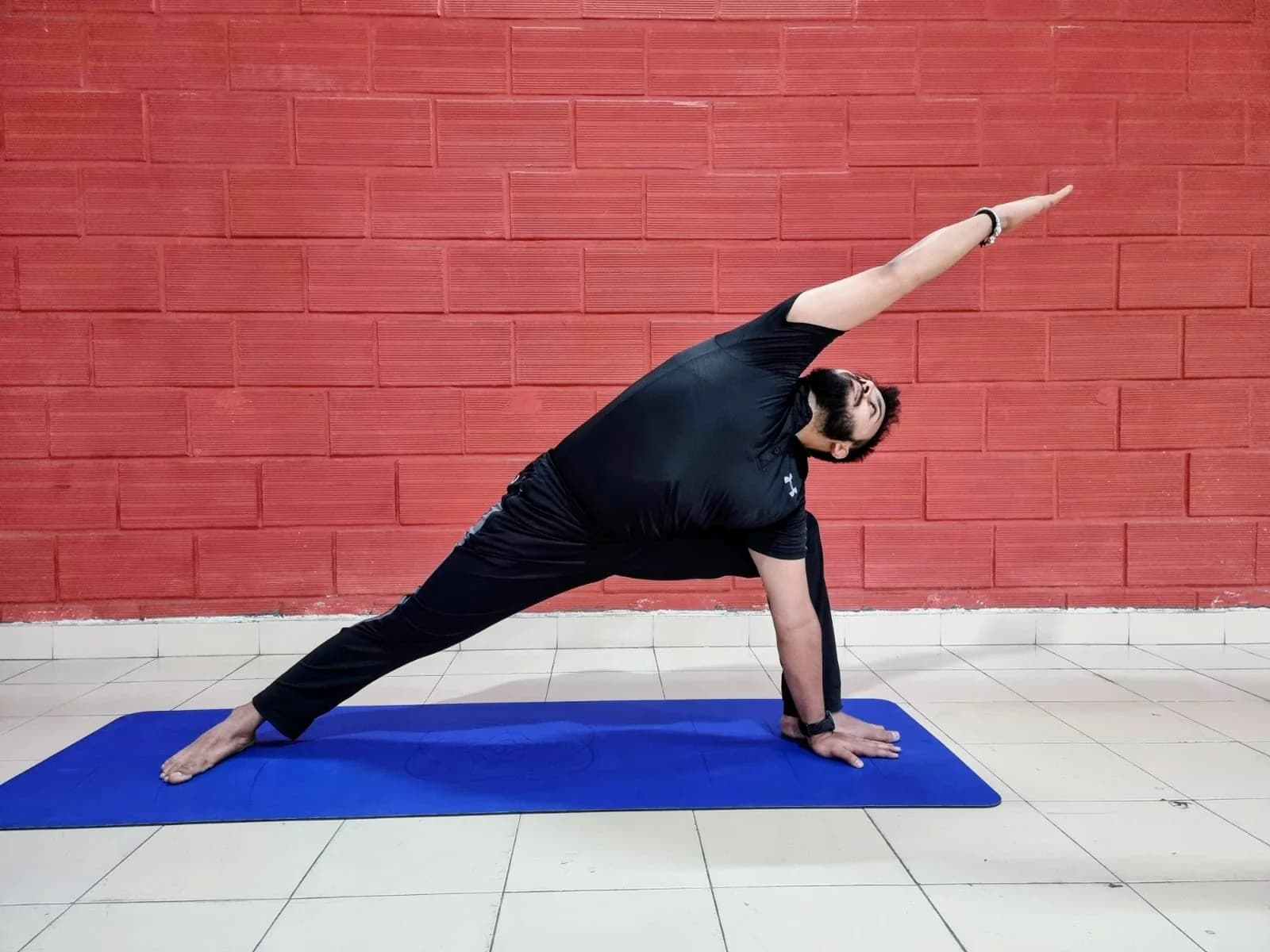 Gandharva in profile holding a side-angle triangle pose against a brick wall